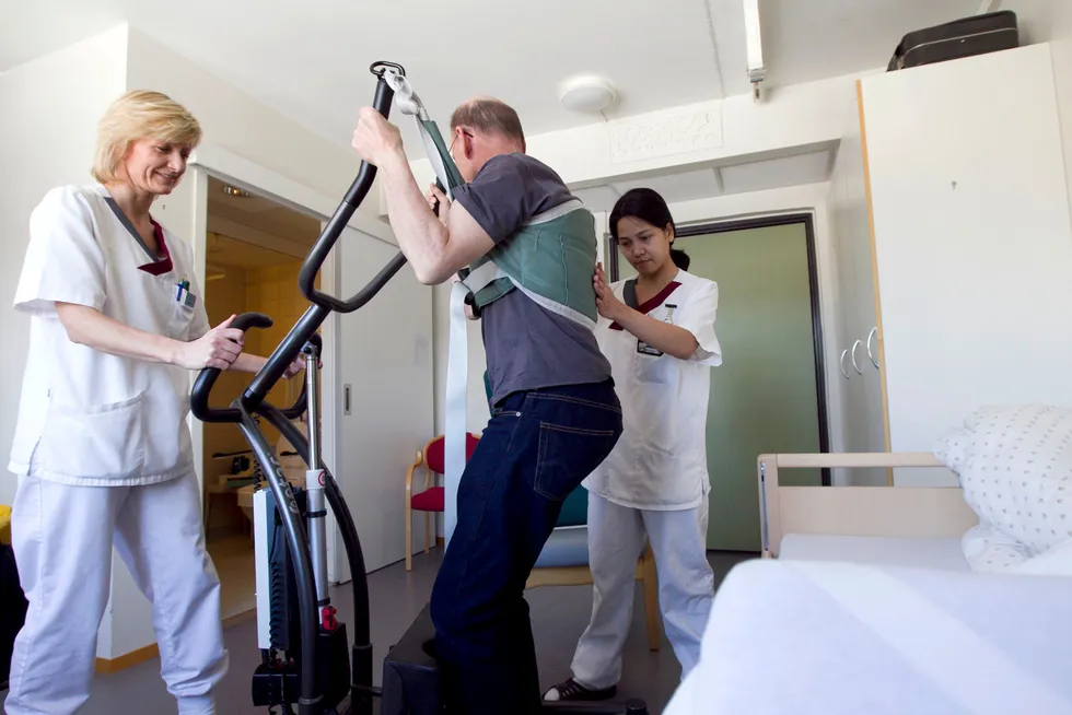 Helsefagarbeidere hjelper en mann opp av sengen med et løfteapparat. Illustrasjonsbilde.