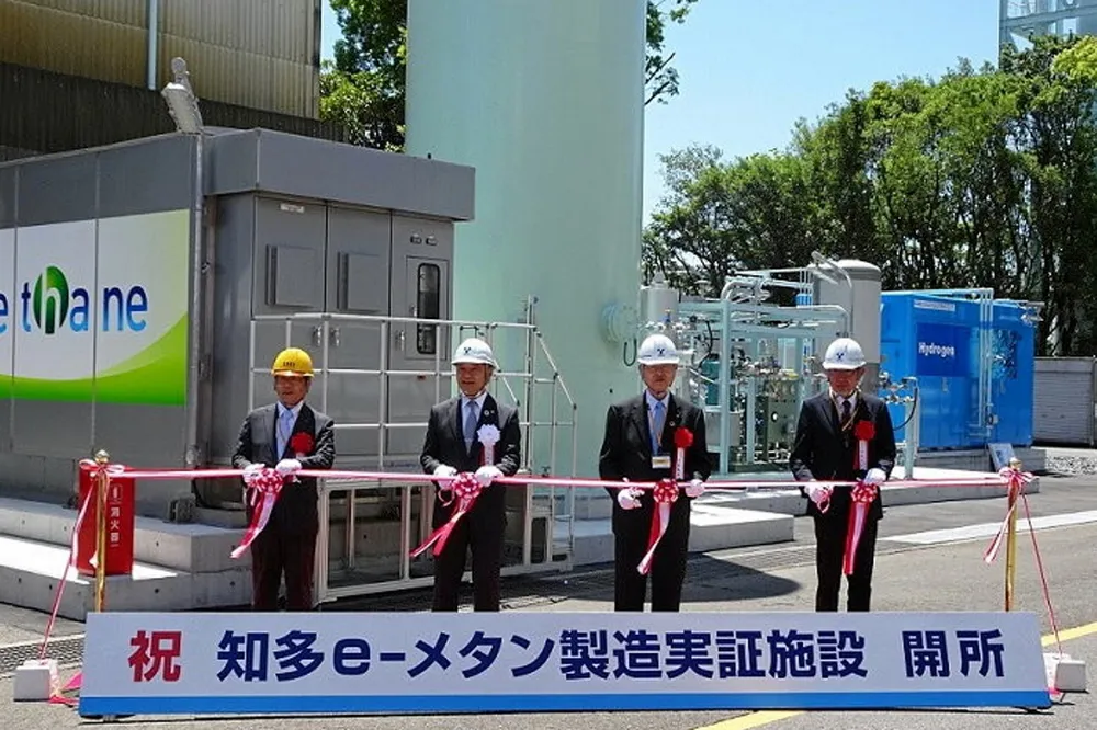 The ribbon-cutting ceremony for the project in Chita, Aichi prefecture. The blue structure on the right is the electrolyser, and the dark grey structure on the left (labelled 'e-methane' in English) contains the Sabatier equipment. The sign at the front reads: 'Congratulations [in red] Chita e-methane production demonstration facility opened'